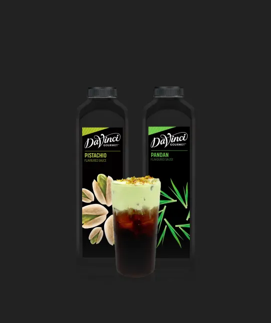 Two DaVinci Gourmet sauce bottles (Pistachio and Pandan) behind a layered iced coffee on a dark background.