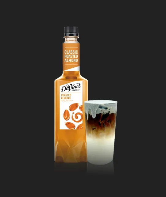 Bottle of DaVinci Gourmet Classic Roasted Almond syrup with a glass of iced coffee beside it on a dark background.