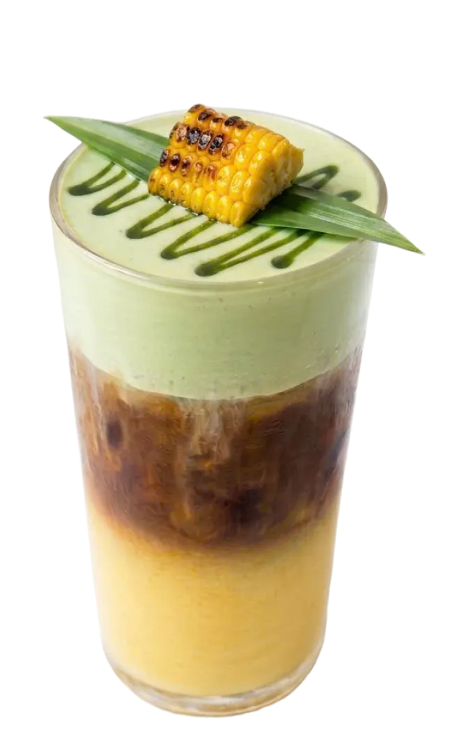 Layered smoothie in a tall glass topped with green mousse, corn cob garnish, and a leaf, drizzled with green sauce on top.